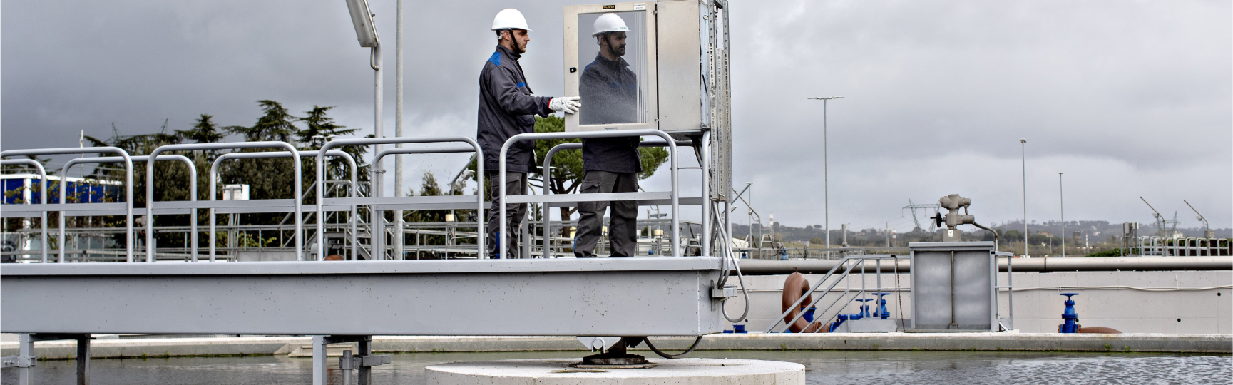 Due operatori Acea Ato 5 con caschi di sicurezza controllano un pannello su una passerella in un impianto industriale