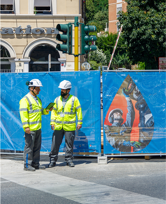 Tecnici in abbigliamento ad alta visibilità e casco bianco parlano davanti a un cantiere cittadino